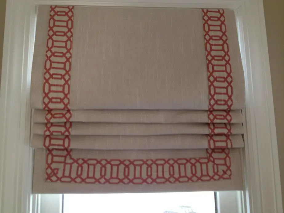 A linen Roman shade with a coral geometric border trim, raised in soft folds inside a white window frame